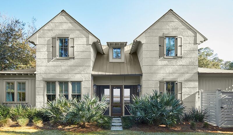Stylish off-white house with gray-green shutters, brown door, breezeway, and greenery.