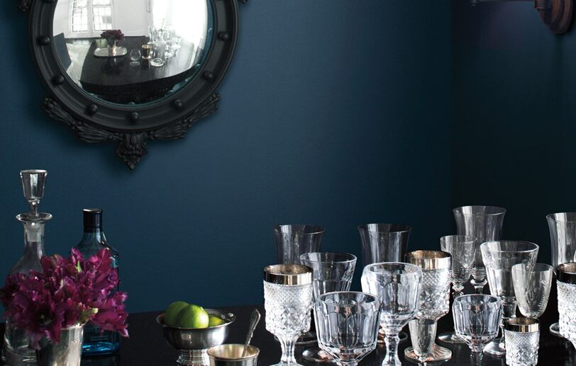A dark blue-painted dining room wall frames ornate glasses on a table. A dark blue-painted dining room wall frames ornate glasses on a table.