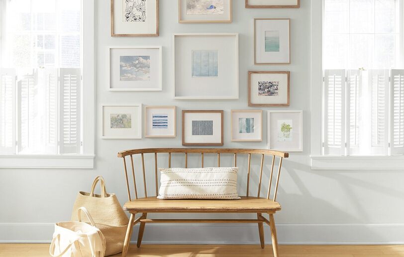 An off-white painted wall with a collection of framed art behind a wooden bench and shopping bags.